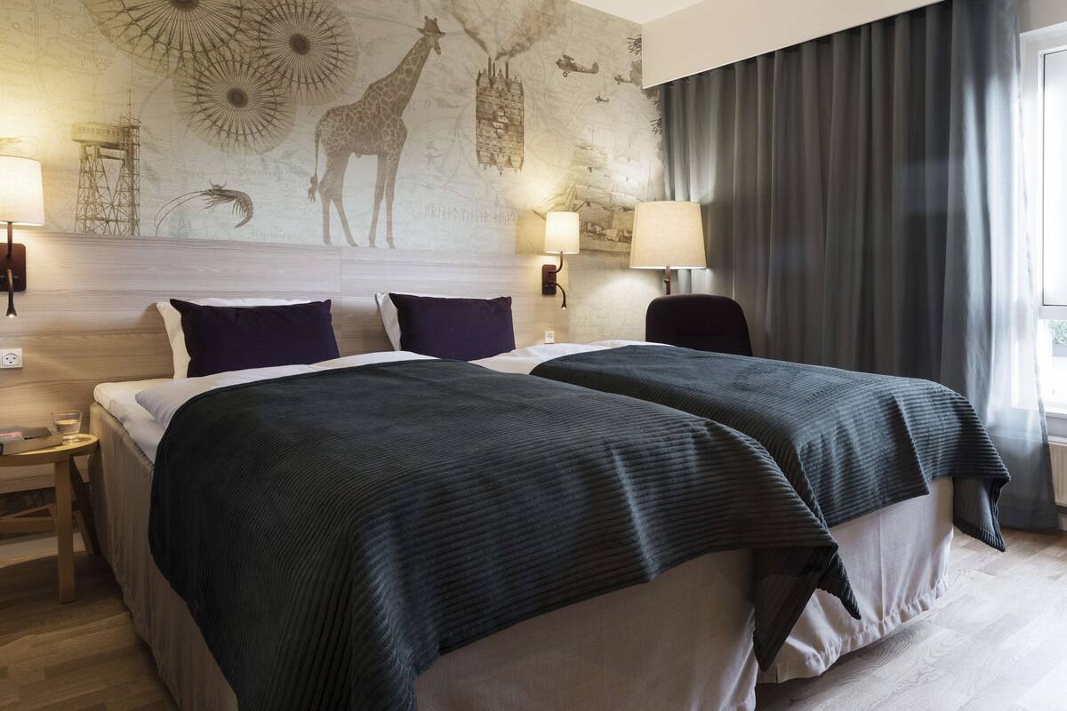 Hotellihuone, jossa on kaksi sänkyä, suuri seinämaalaus ja näkymä ulos.