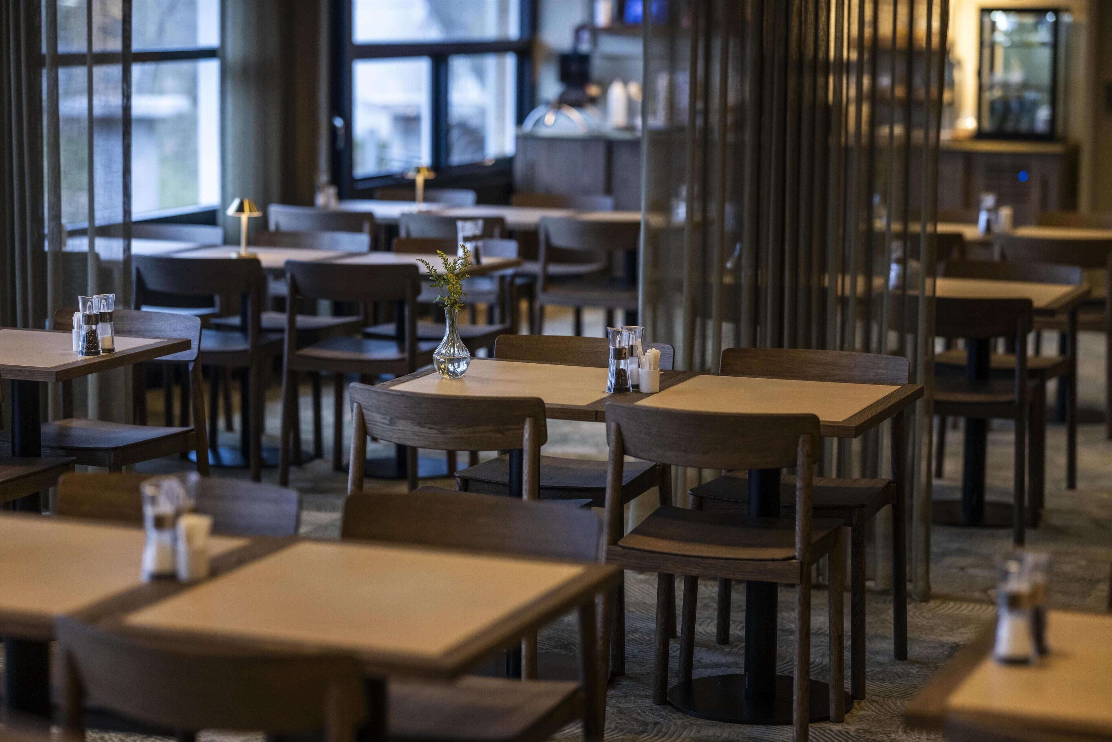 L’intérieur d’un restaurant, avec des tables et des chaises en bois, un vase rempli de fleurs et une grande fenêtre en arrière-plan.