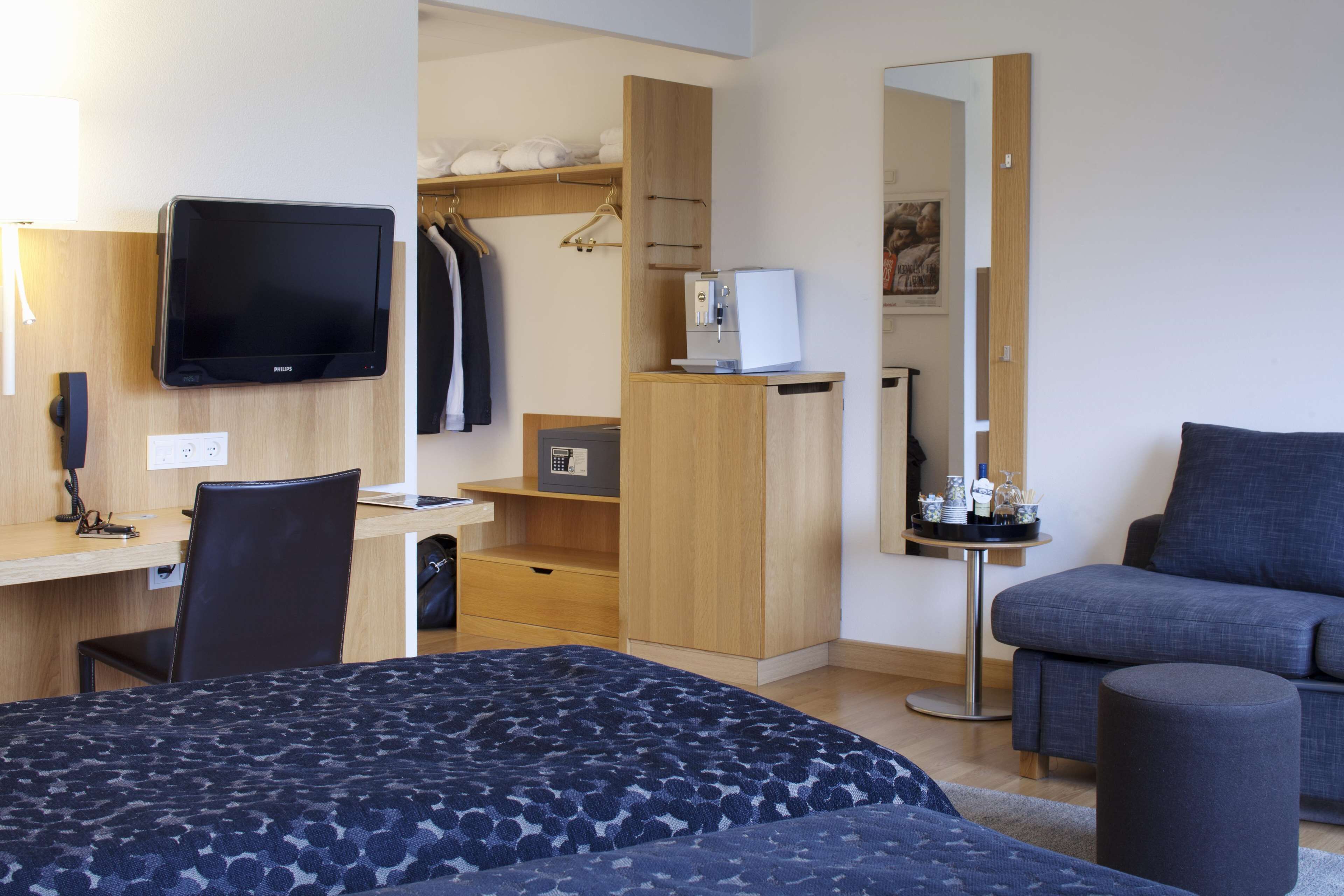 Habitación de hotel con cama, escritorio con computadora, televisor, armario y zona de estar con sofá y reposapiés.
