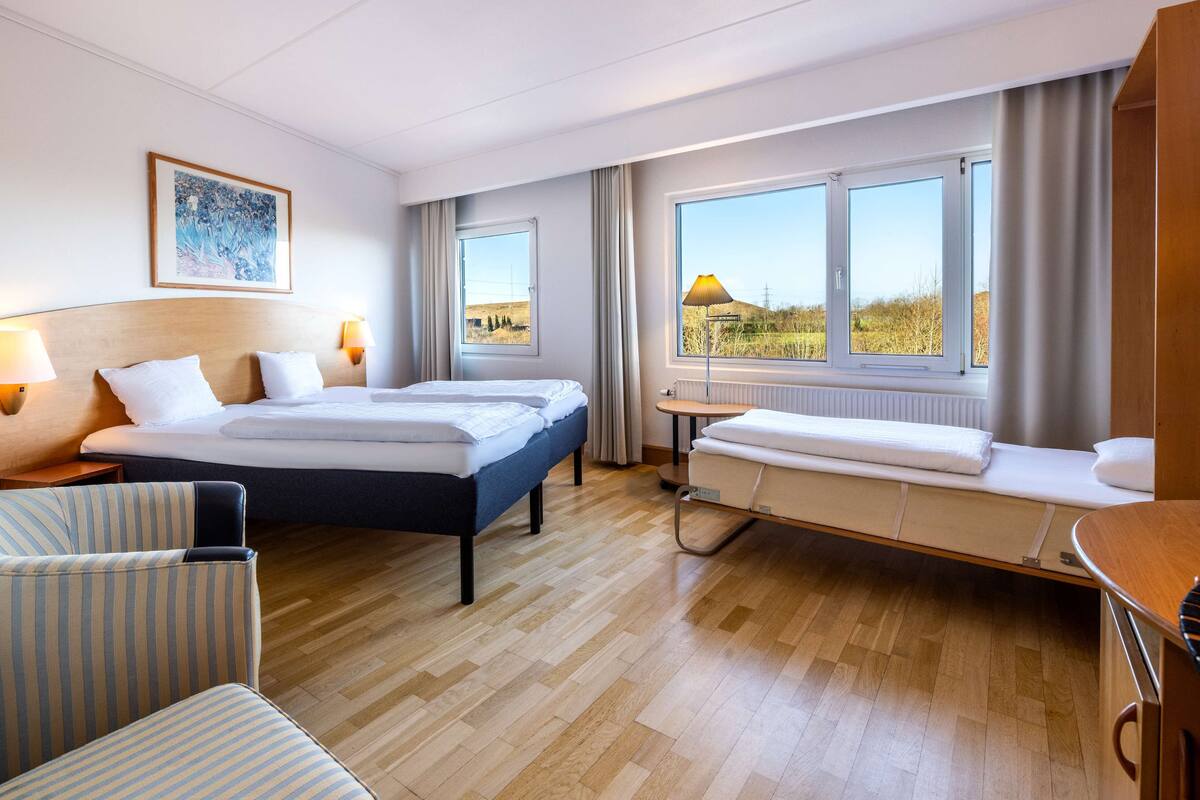 A hotel room with two beds, a desk, a chair, and large windows with a view of the countryside.