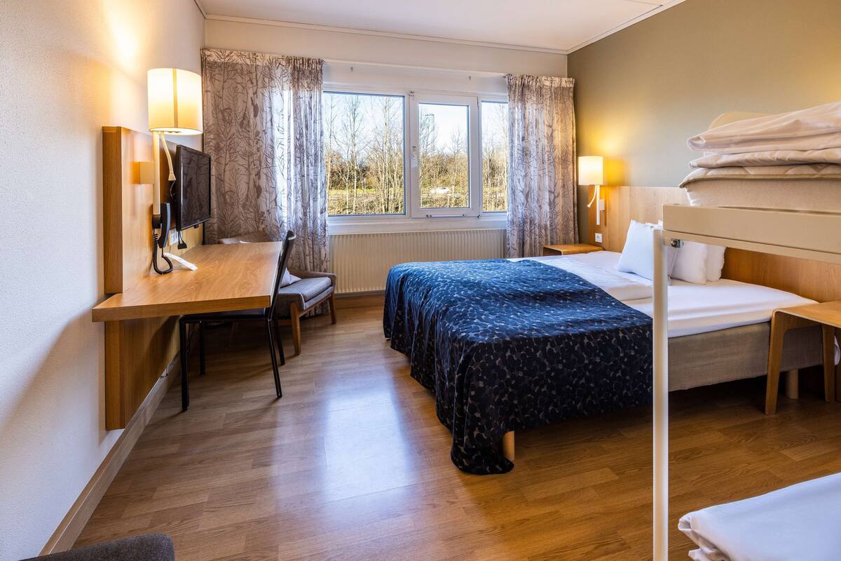 A hotel room with a wooden desk, a bed with a dark patterned bedspread, a bunk bed, and a window with curtains.