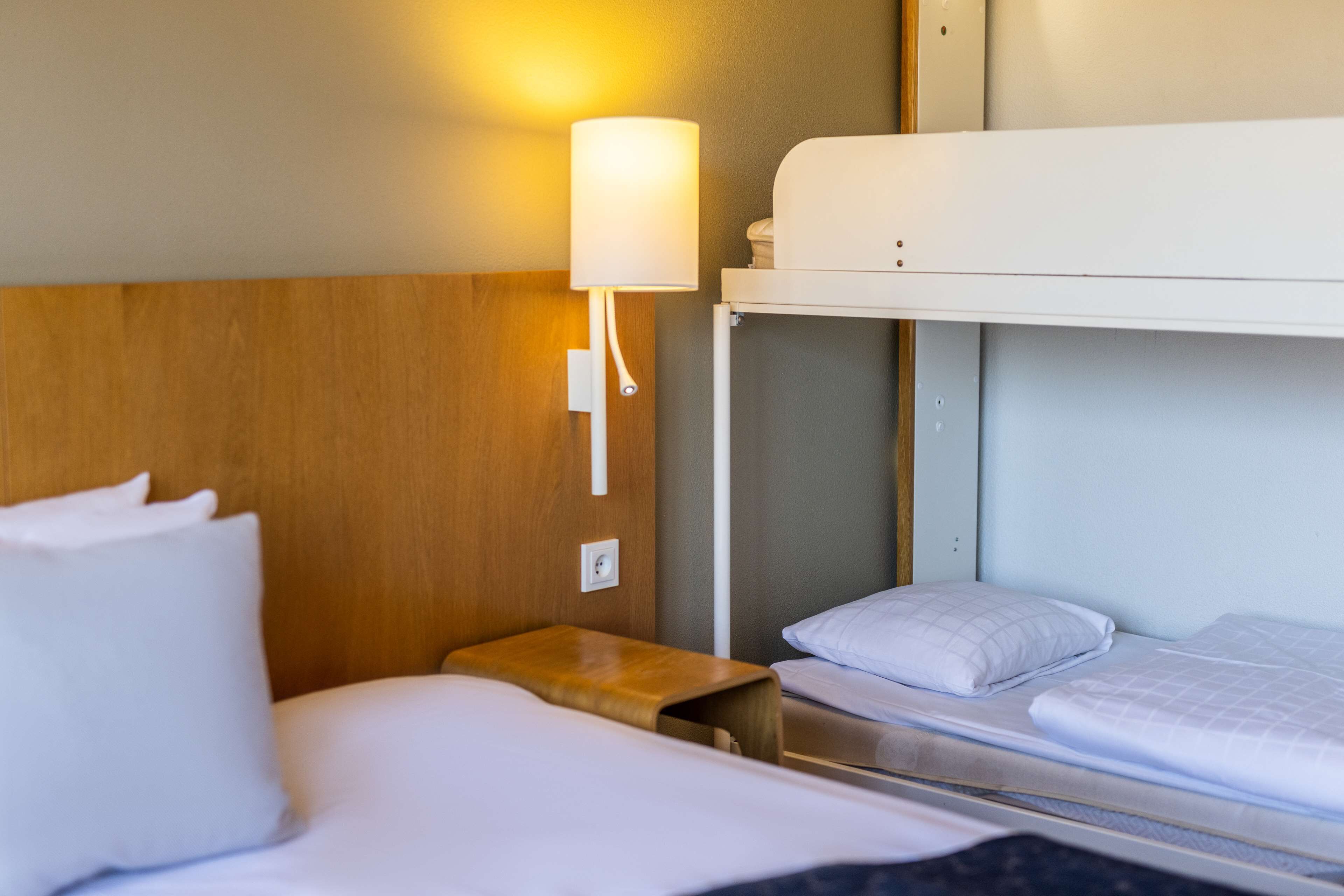 Une chambre d’hôtel avec un lit superposé, une table de chevet, une lampe et une prise électrique au mur.