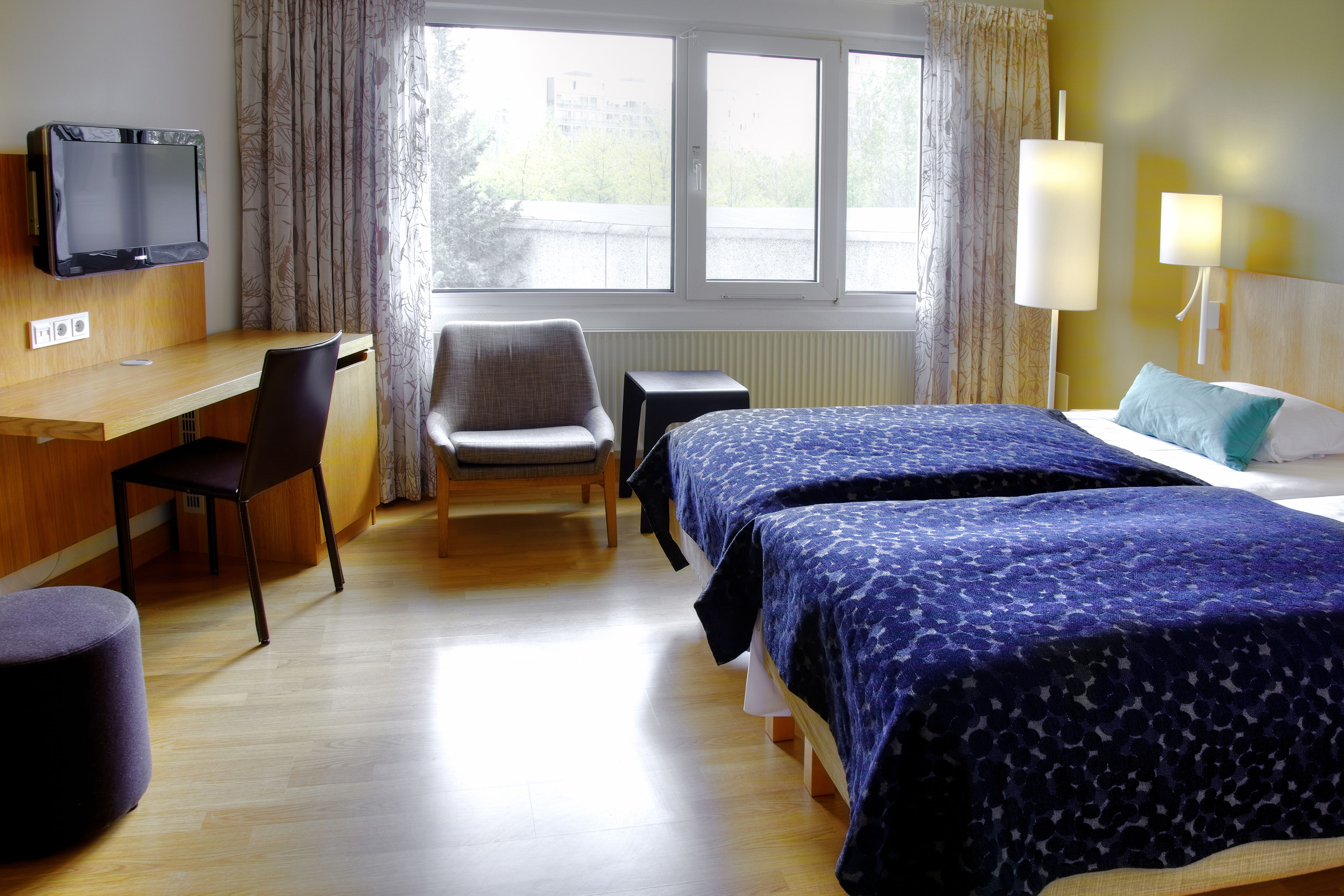 Et hotelværelse med et træskrivebord, en stol, en seng med blåt sengetøj, et fjernsyn monteret på væggen og et vindue med gardiner.