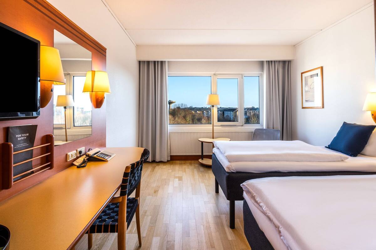 A hotel room with a desk, two beds, a TV, and a window with a city view.