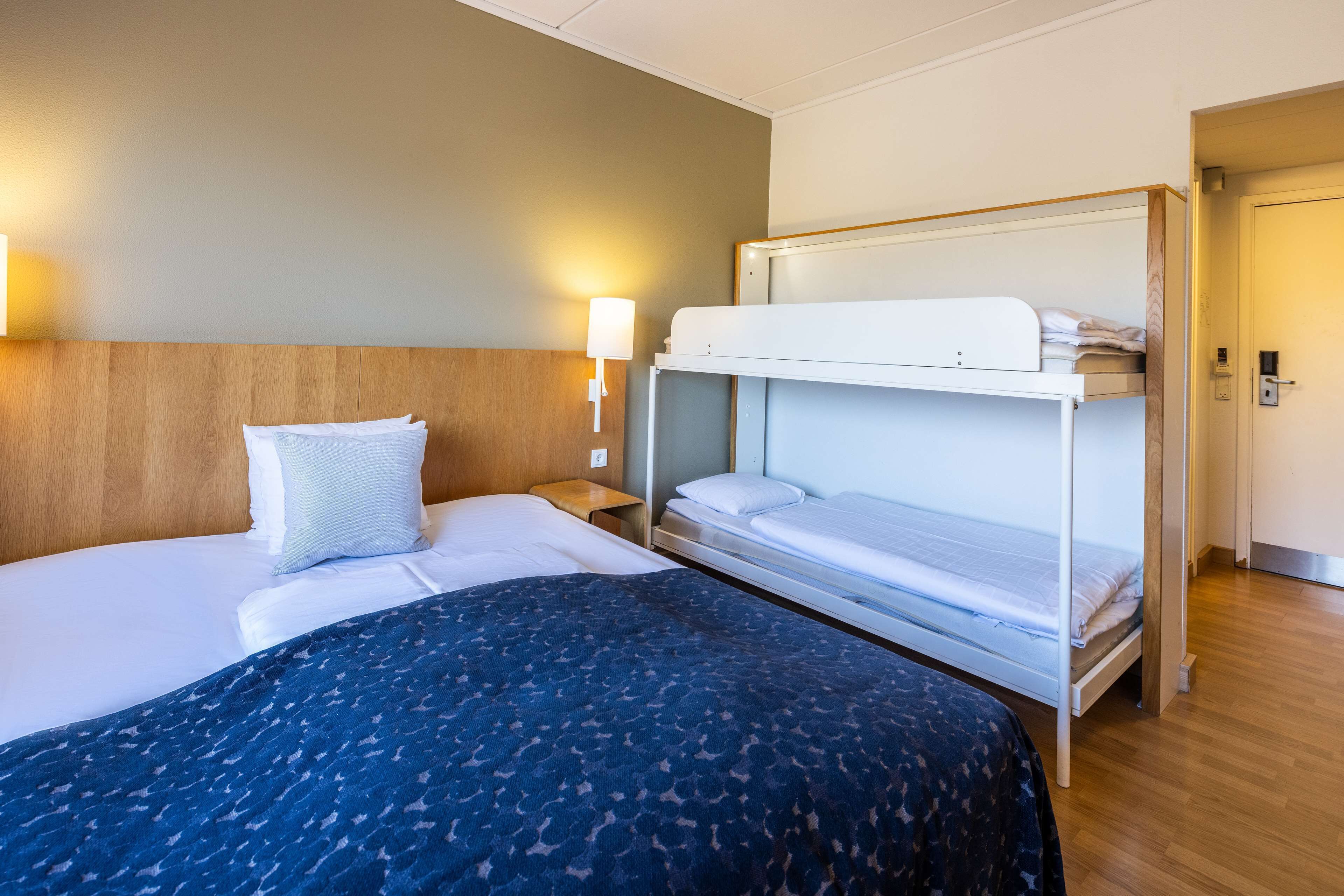 Una habitación de hotel con una cama individual, una cama literera, una lámpara de noche y un cabecero de madera.