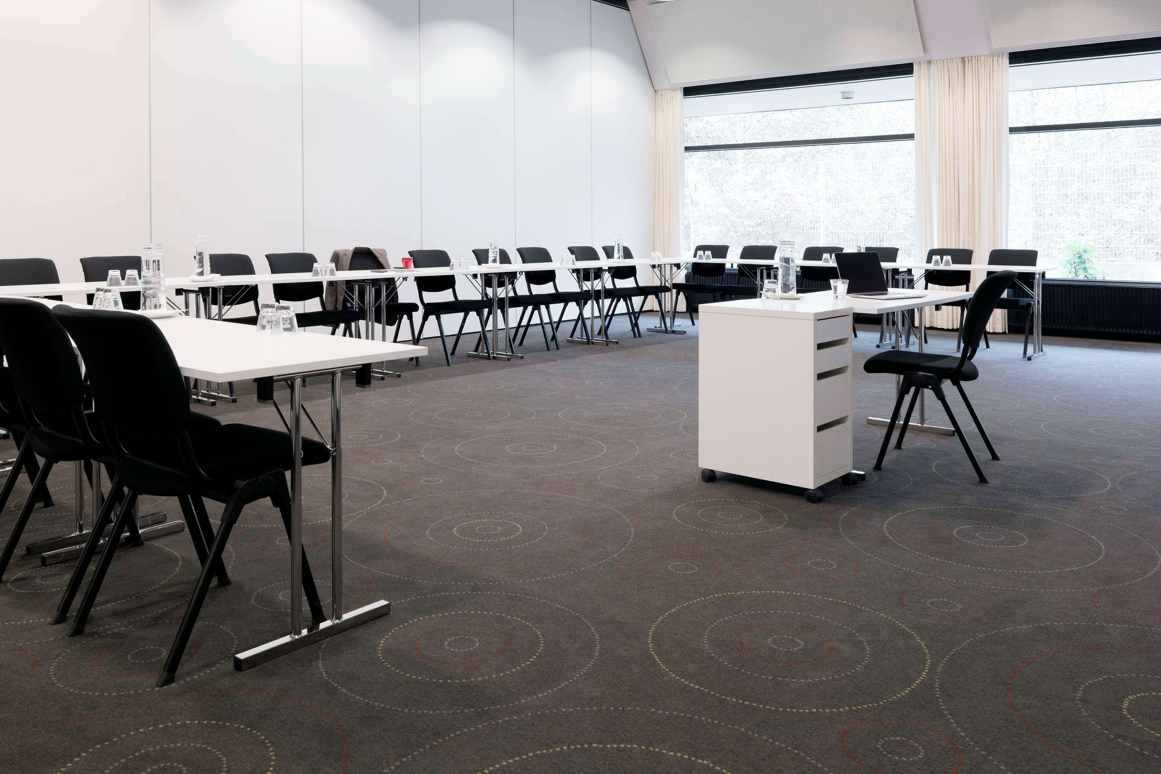 Een vergaderzaal met rijen zwarte stoelen, witte tafels en een wit podium.