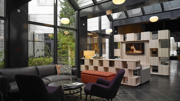 A modern living room with a black leather sofa, a red ottoman, and a black coffee table. There are two purple armchairs, a glass of water, and a magazine on the table. The room has large windows, a black floor, and a wooden shelving unit with a television on top. - Scandic Aarhus Vest (Aarhus)