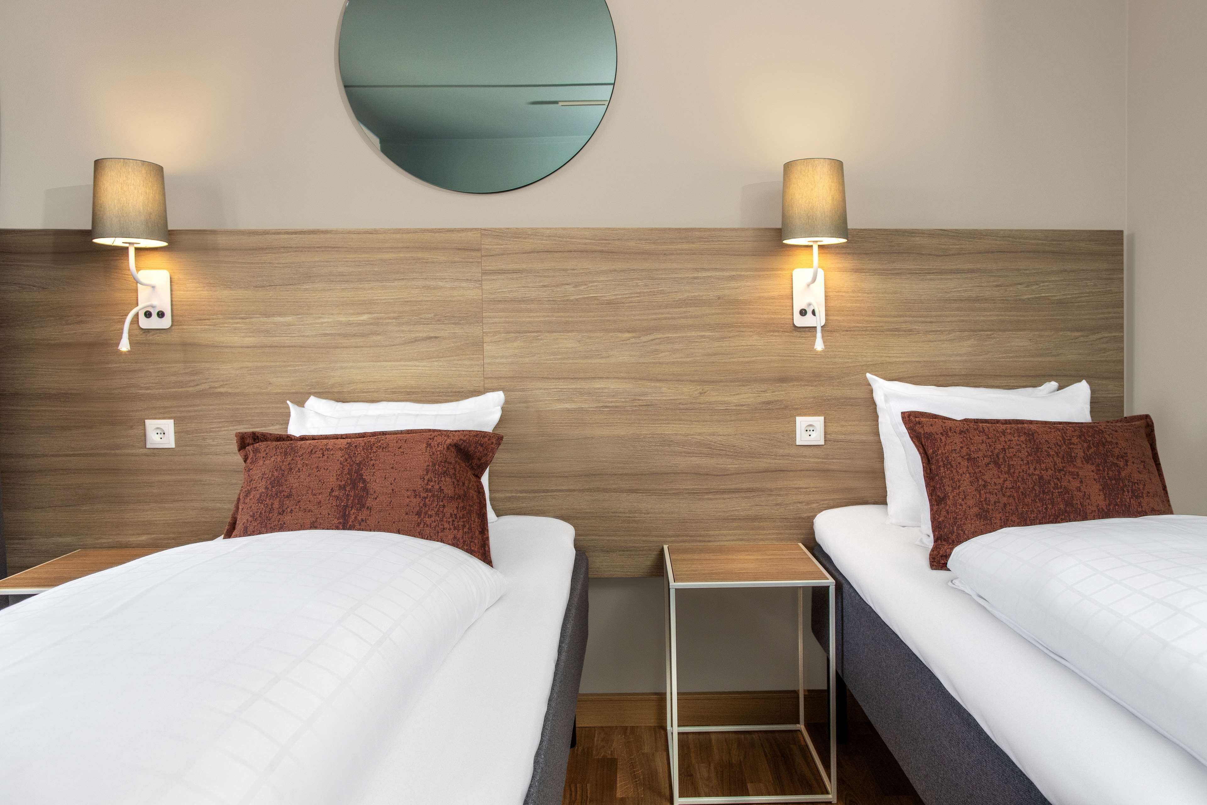 Habitación de hotel con dos camas, cada una con ropa blanca y almohadas marrones, cabeceros de madera y lámparas fijadas a la pared.