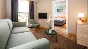 A modern hotel room with a sectional sofa, a green armchair, a small round table with a vase of flowers, a flat-screen TV, and a bed with orange pillows. - Scandic Roskilde Park (Roskilde)