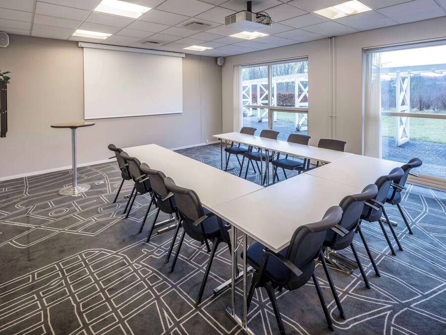 Un salón de conferencias con una larga mesa, sillas, un proyector, una pizarra y ventanas grandes.