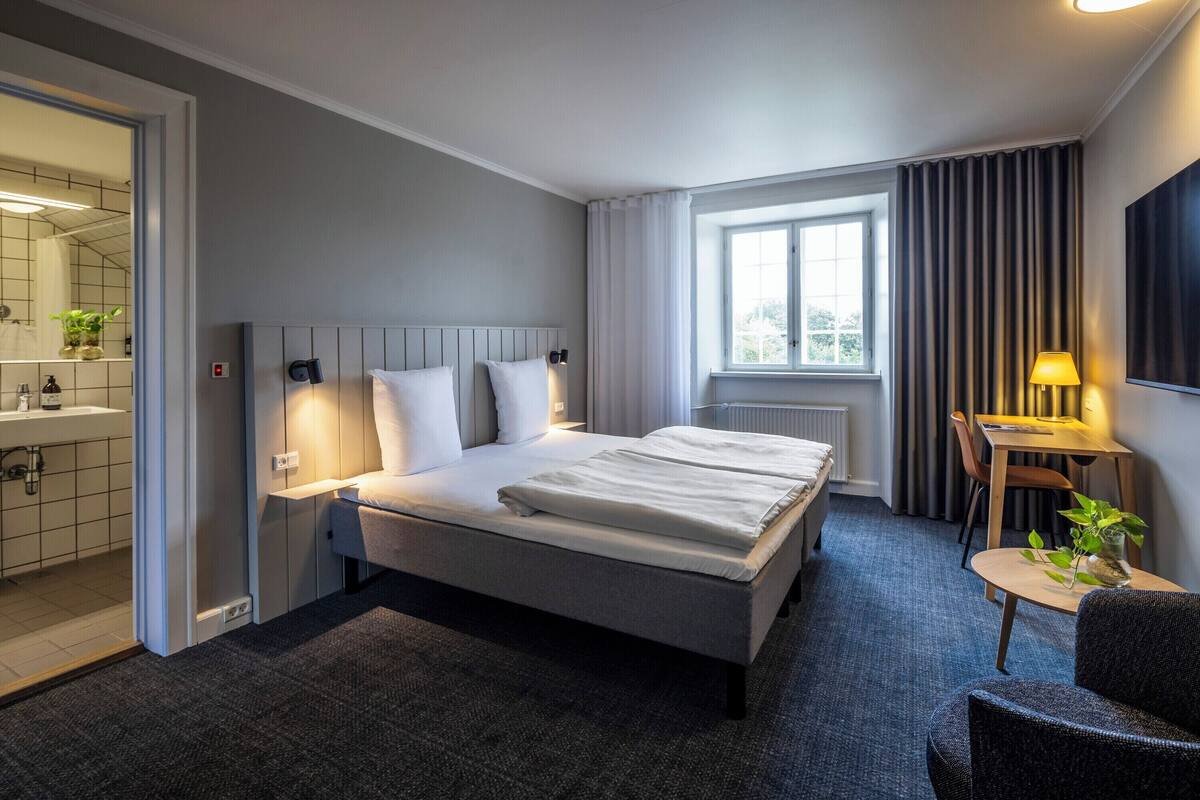 A modern hotel room with a large bed, a bedside table with a lamp, a small table with a plant, a window with curtains, and a bathroom visible through an open door.
