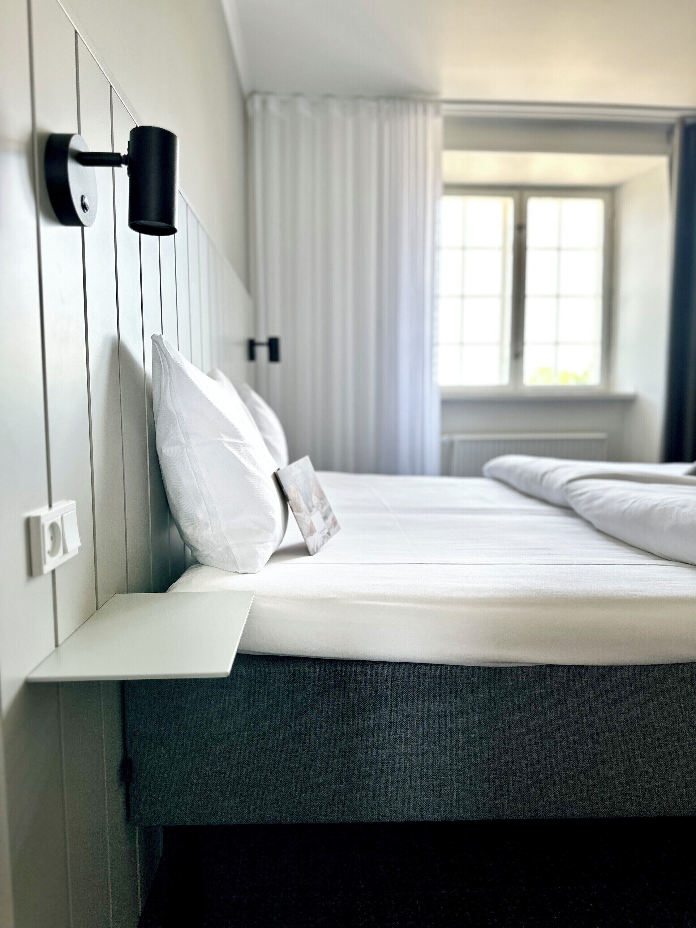 A modern hotel room with a neatly made bed, a bedside table, and a wall-mounted lamp.