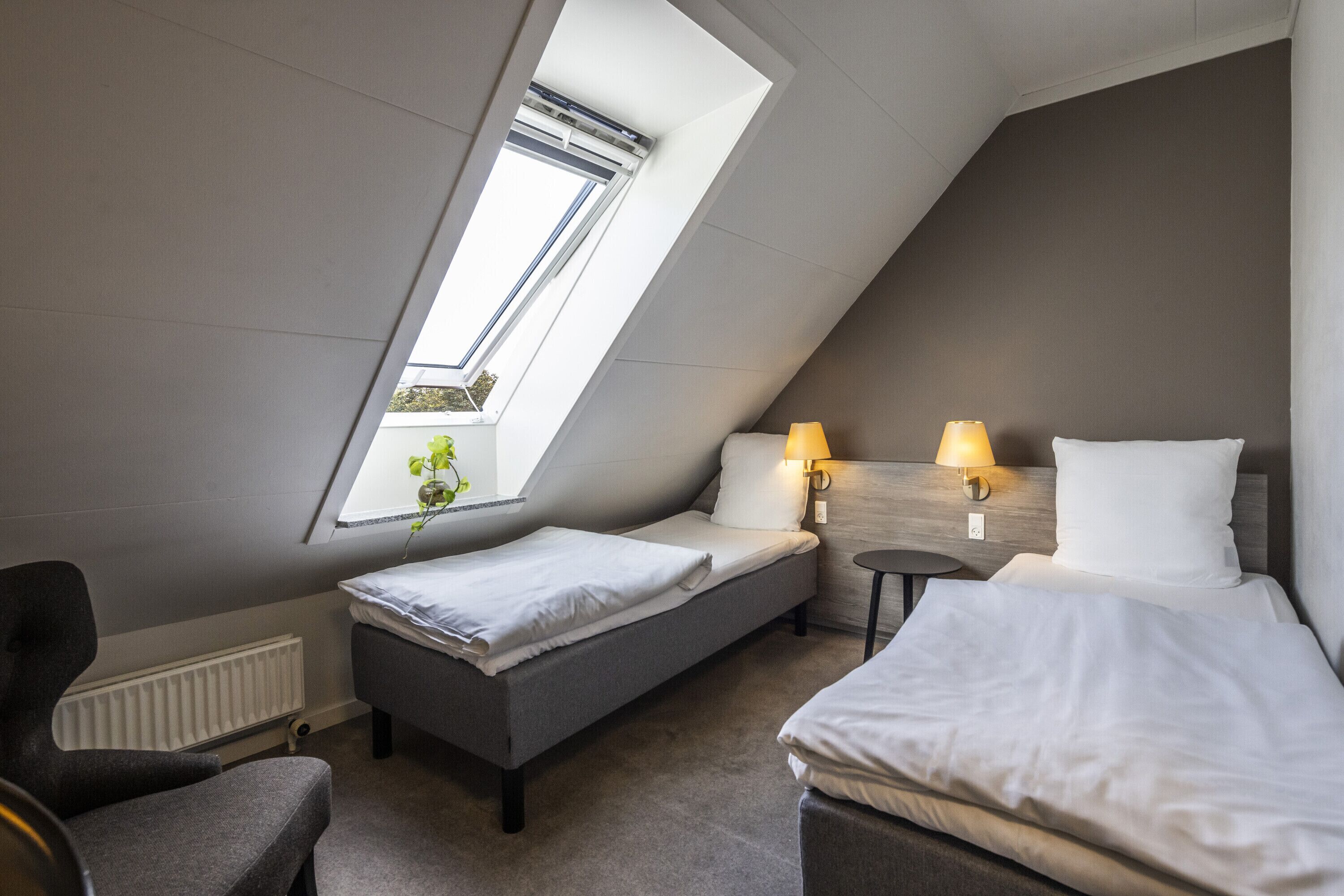 A modern hotel room with a sloped ceiling, two single beds, a small table with lamps, a radiator, and a window with a plant.