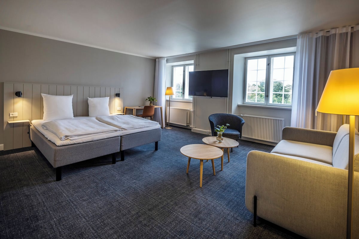 A hotel room with a bed, two bedside tables, a sofa, a TV, and a window with curtains.