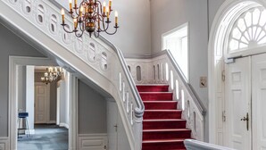 A grand staircase with red carpet and white balustrades, leading to a room with a chandelier and a large arched window. - Comwell Bygholm Park (Horsens)