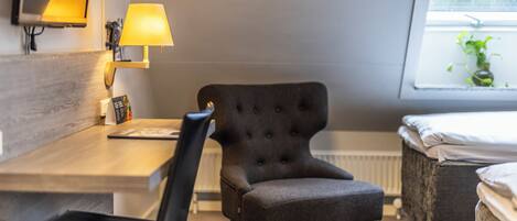 A hotel room with a wooden desk, a black armchair, a bedside table, a lamp, and a window with a view of greenery.