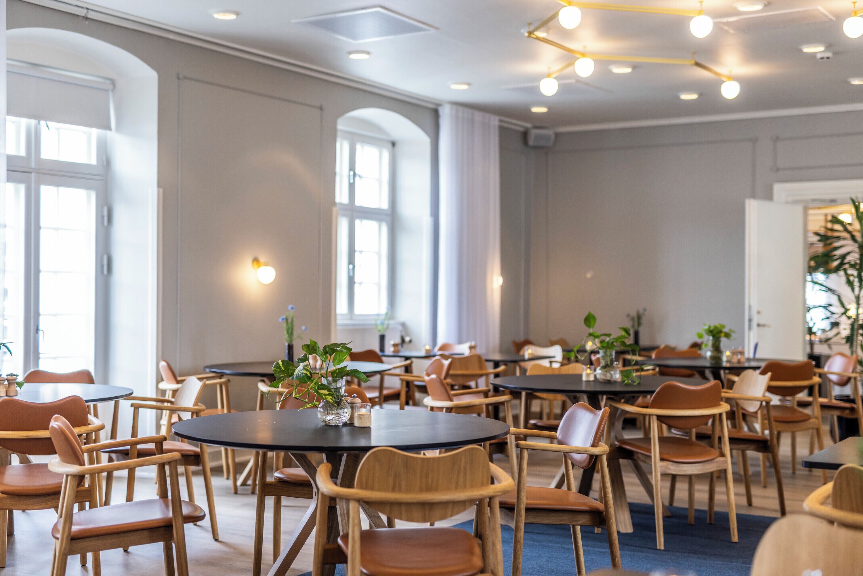 A spacious dining area with round tables and wooden chairs, large windows, and ceiling lights.