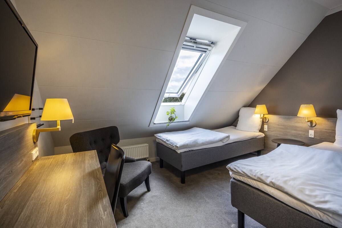 A modern, compact room with a skylight, two beds, a desk, and two lamps.