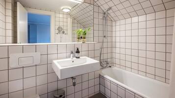 A bathroom with a white sink, mirror, and bathtub. There are potted plants on the sink and a trash can in the corner.