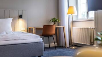 Habitación de hotel con cama, escritorio, silla, lámpara, planta y ventana con cortinas.