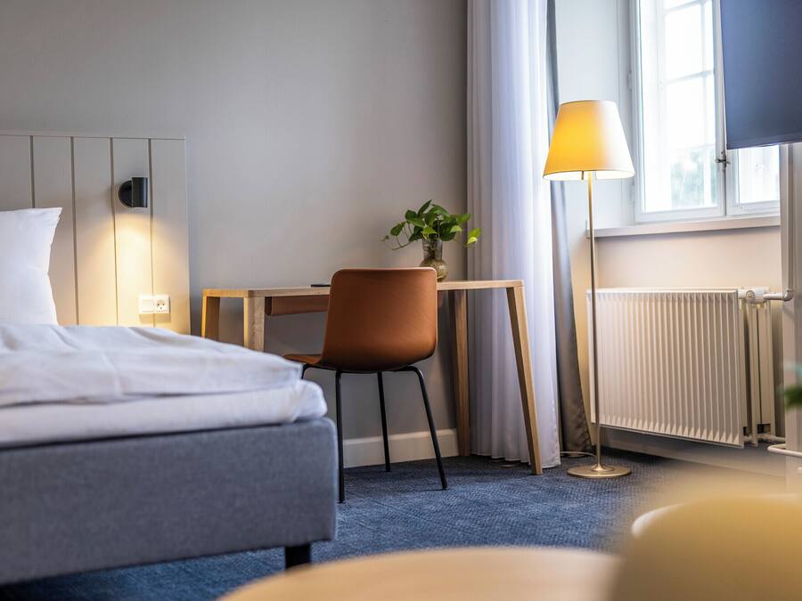 Habitación de hotel con cama, escritorio, silla, lámpara, planta y ventana con cortinas.
