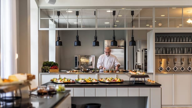 A modern kitchen with a chef preparing food, a buffet counter with various dishes, and a wine display. - Comwell Bygholm Park (Horsens)