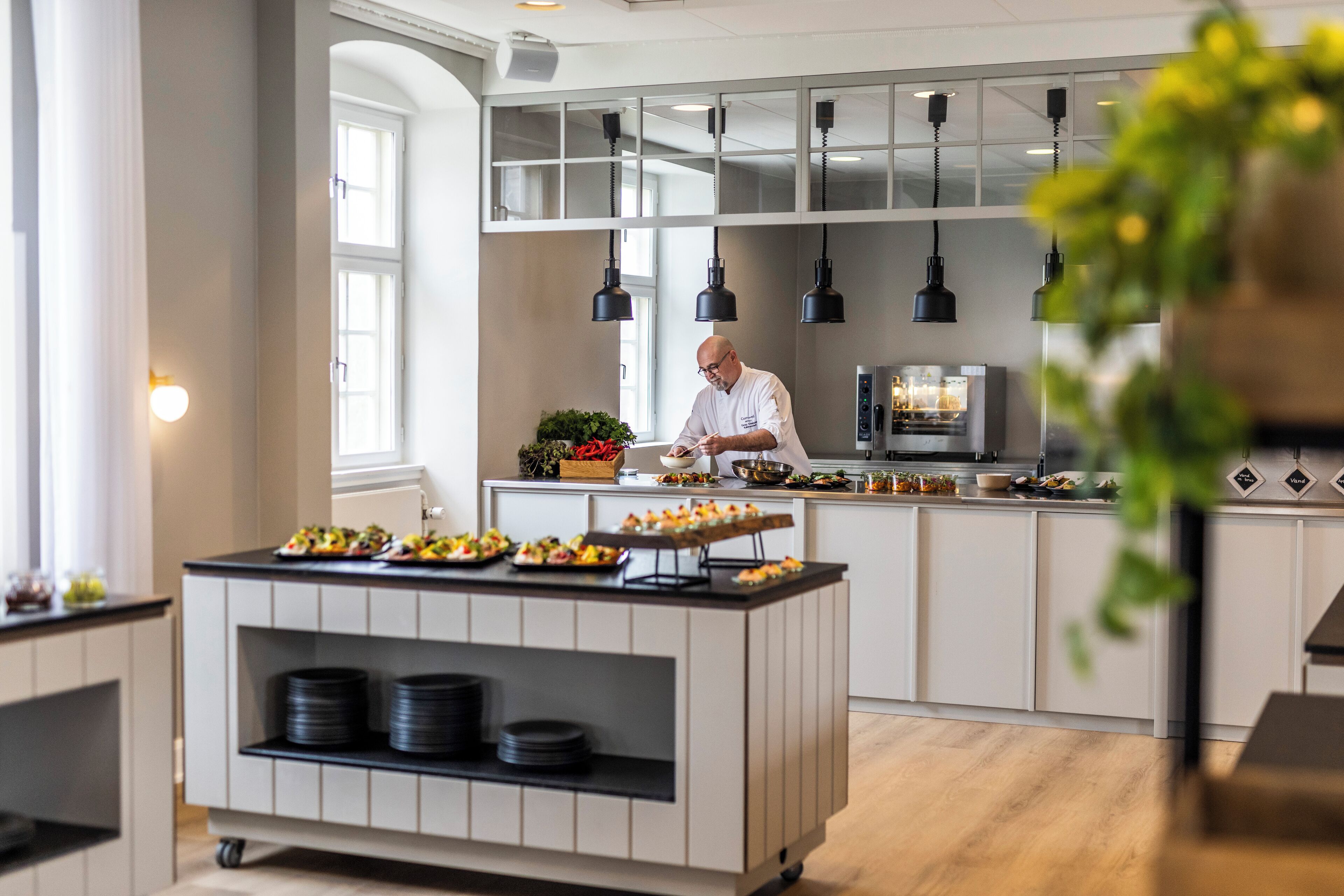 Una cocina moderna con un chef preparando comida, una estación de buffet con platos y un refrigerador al fondo.