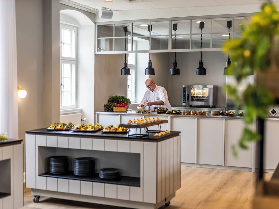 Una cocina moderna con un chef preparando comida, una estación de buffet con platos y un refrigerador al fondo.