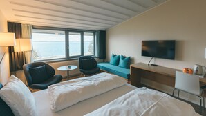 A hotel room with a bed, two chairs, a desk, a television, and a view of the sea.