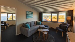 A modern living room with a grey sofa, teal cushions, a coffee table, and a view of the sea through large windows.