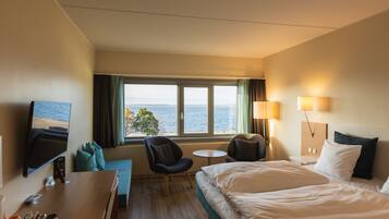 A hotel room with a bed, a desk, a chair, a small table, and a TV. There is a window with a view of the sea.