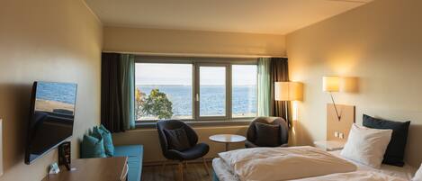 Ein Hotelzimmer mit Bett, Schreibtisch, Stuhl, kleinem Tisch und Fernseher. Ein Fenster bietet Blick aufs Meer.