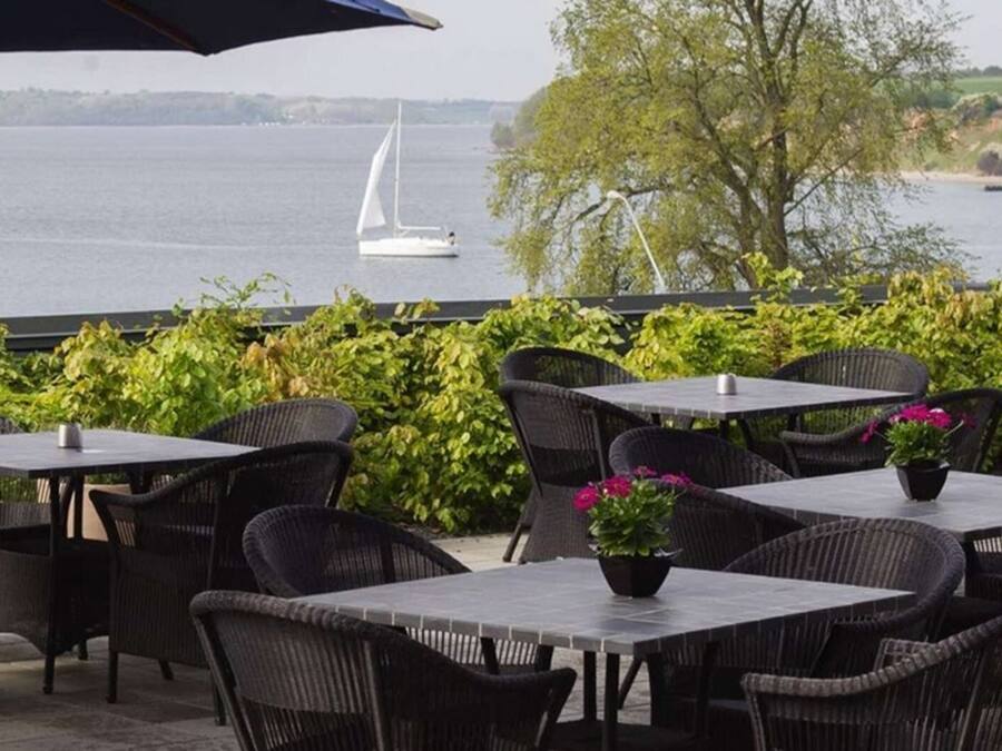 An outdoor patio with tables and chairs, overlooking a lake with a sailboat.