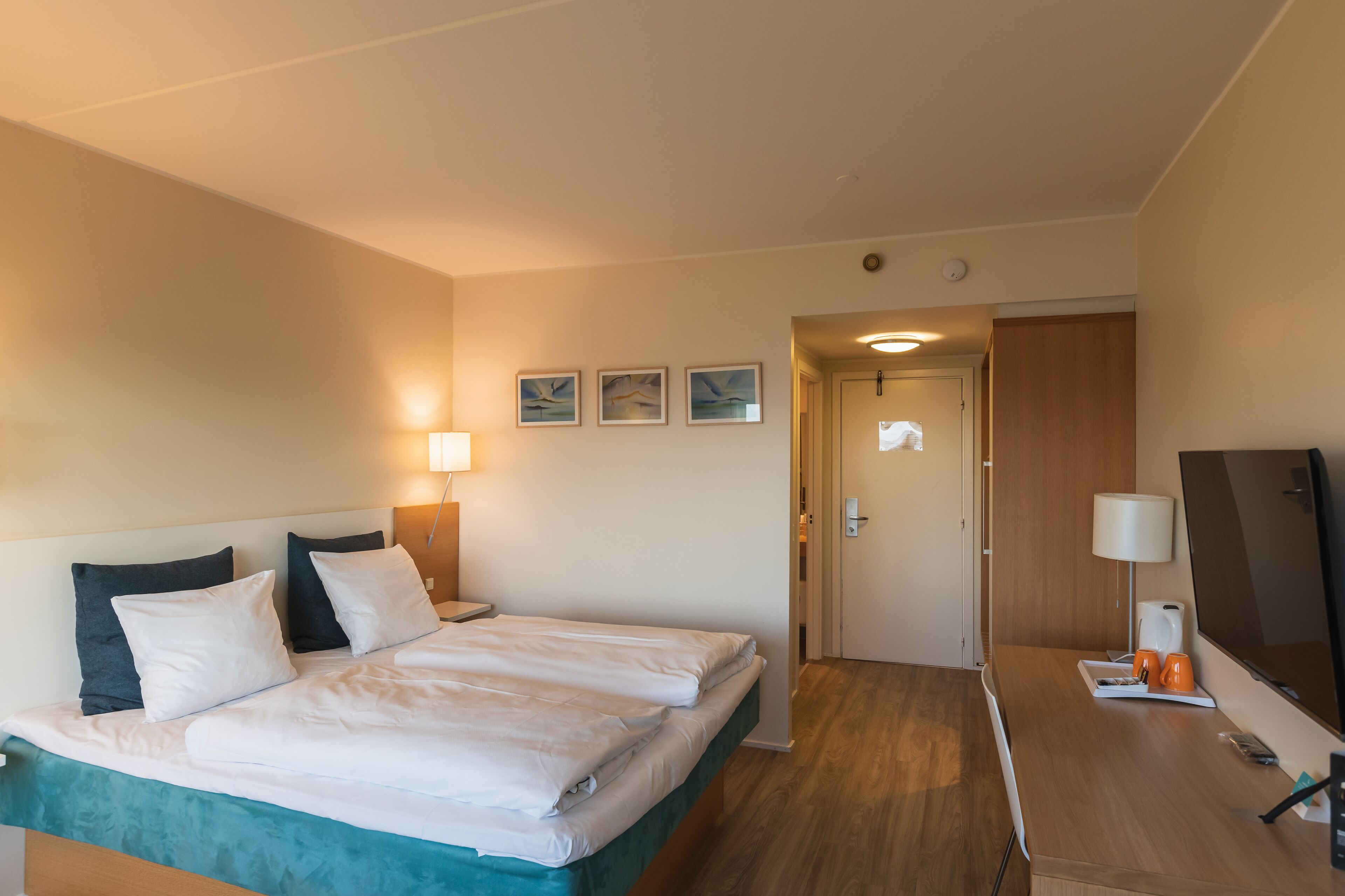 Quarto de hotel com uma cama, mesas de cabeceira, uma escrivaninha com cadeira, televisão e uma porta que dá para outro aposento.