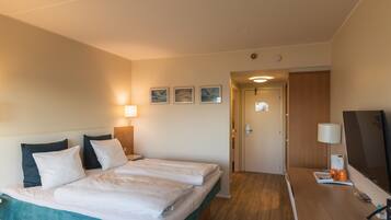 Et hotelværelse med seng, natborde, et skrivebord med stol, et fjernsyn og en dør til et andet rum.