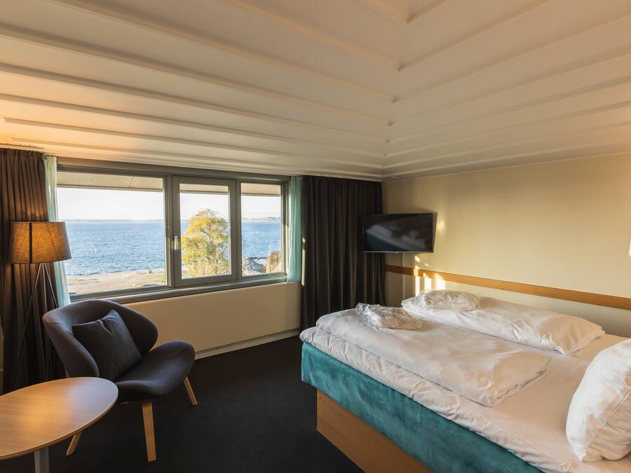 A hotel room with a bed, a chair, a TV, and a window with a view of the sea.