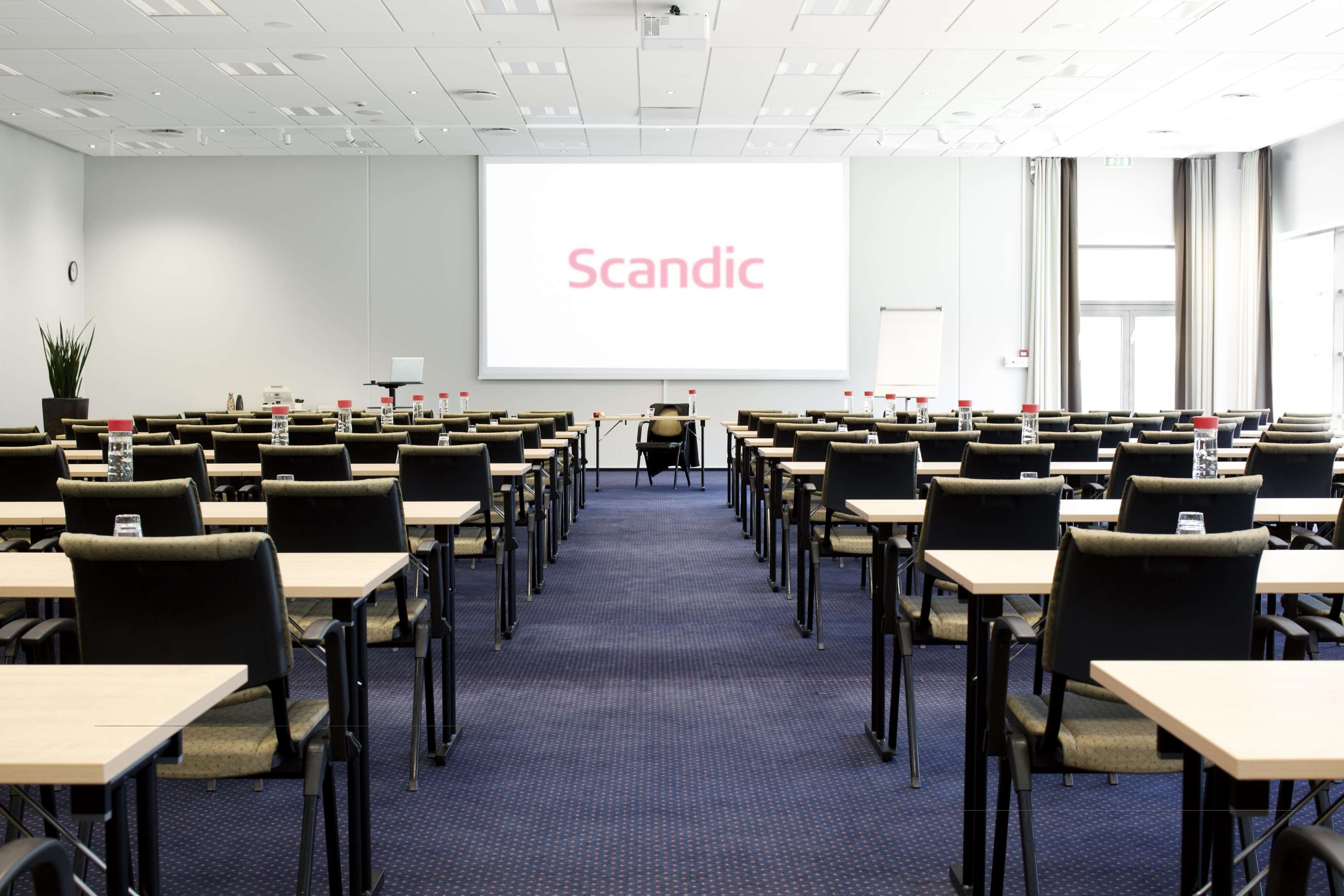 A conference room with rows of tables and chairs, a large screen displaying the word "Scandic," and a potted plant in the corner.