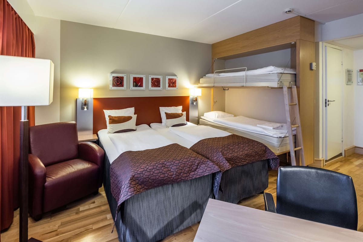 A hotel room with a bed, two bedside tables, a chair, a desk, a bunk bed, and a red curtain.
