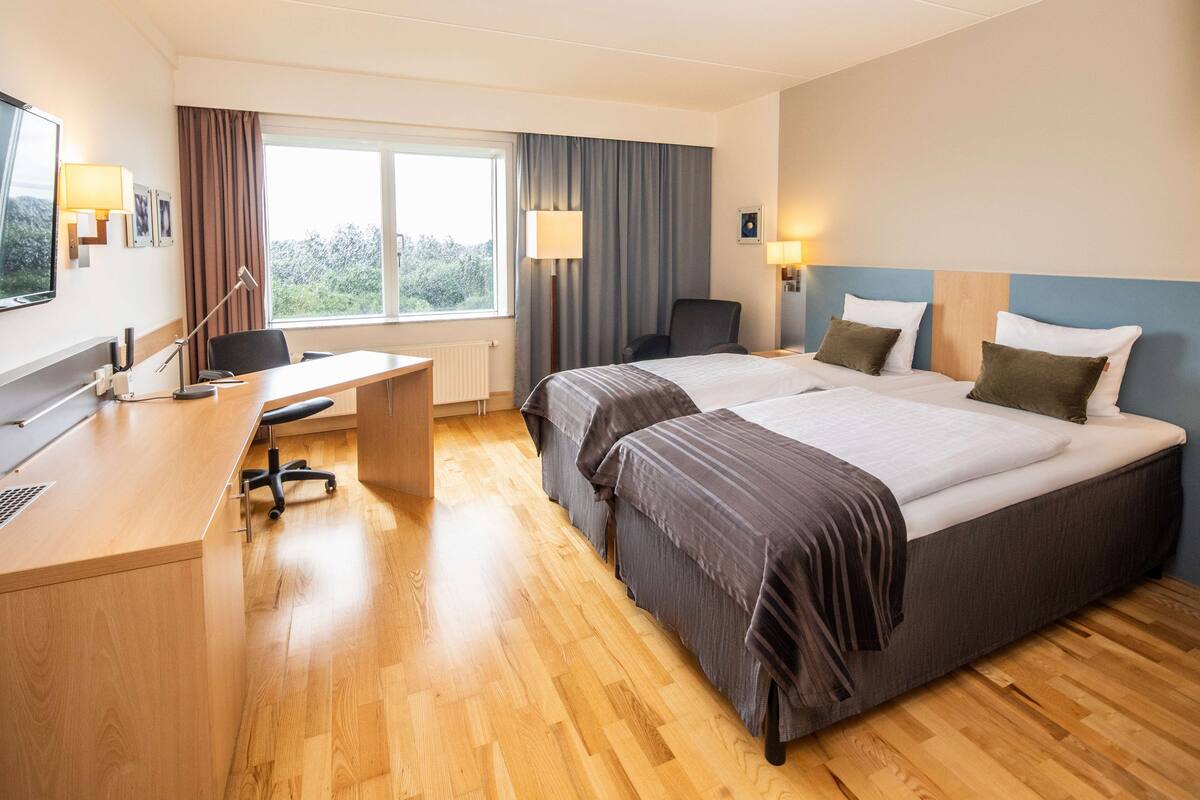 A hotel room with two beds, a desk, a chair, a TV, and a window with curtains.