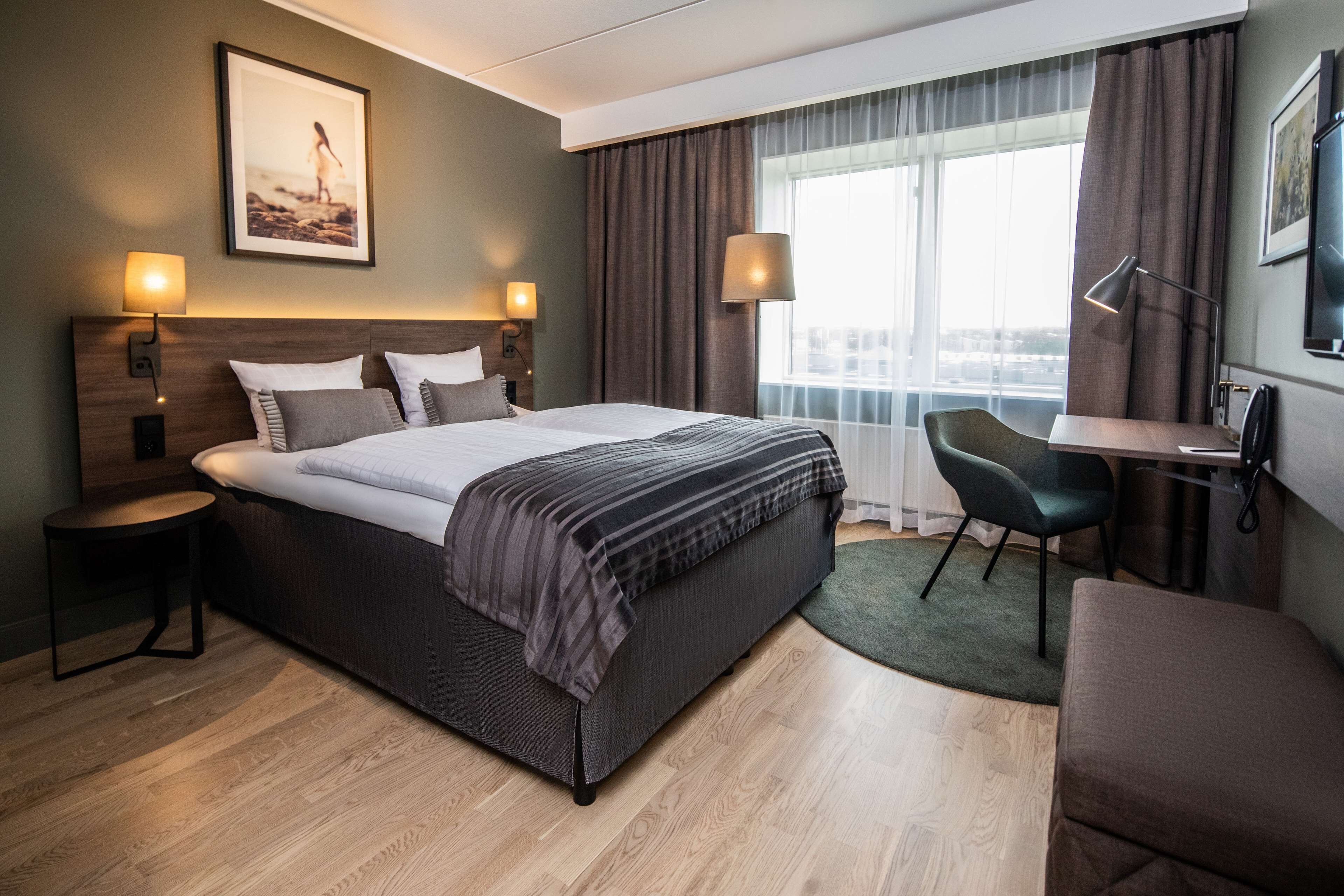 Ett hotellrum med en stor säng, två sängbord, ett skrivbord med en stol, en tv och ett fönster med gardiner.