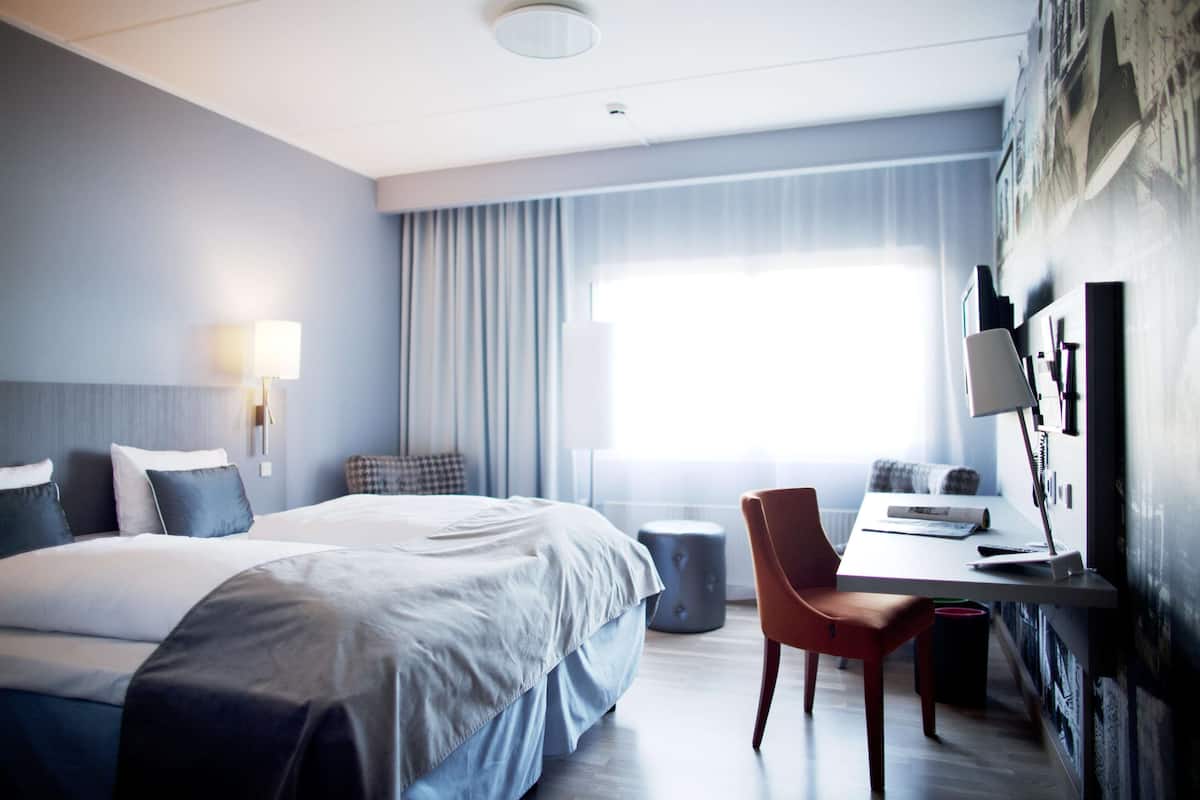 A modern hotel room with a large bed, a desk with a chair, a lamp, and a view of the outdoors through a window.