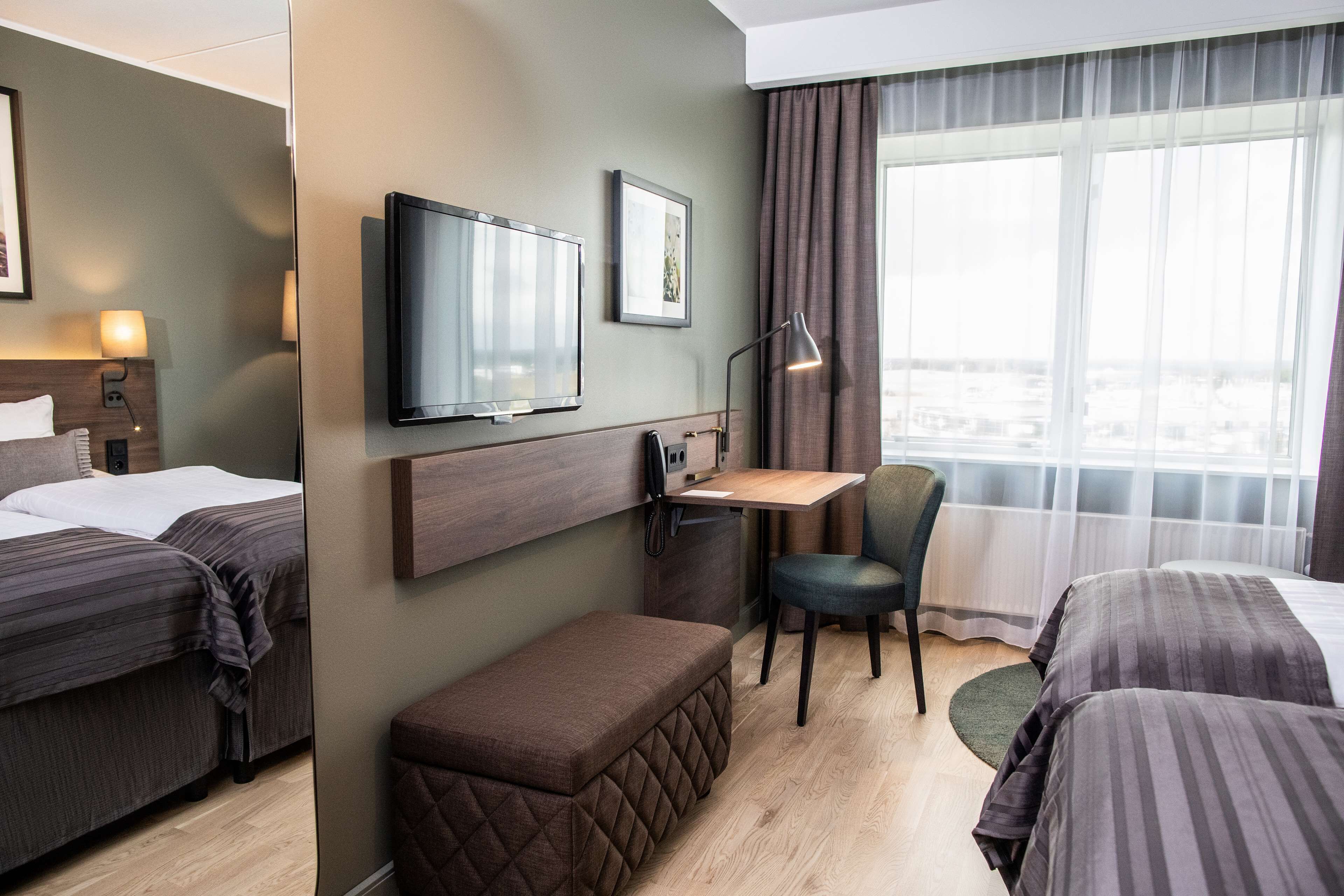 Quarto de hotel com duas camas, uma escrivaninha, uma cadeira, uma televisão e uma janela com cortinas.