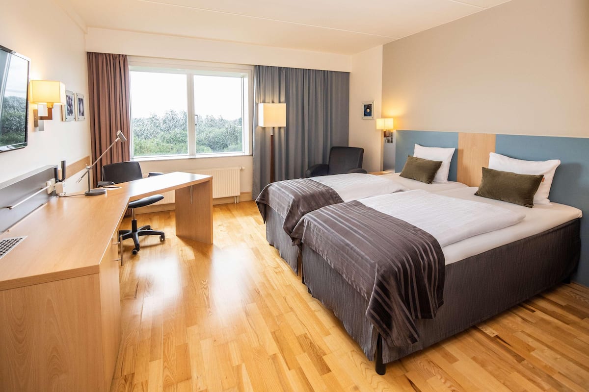 A hotel room with a desk, two beds, a chair, and a window with curtains.