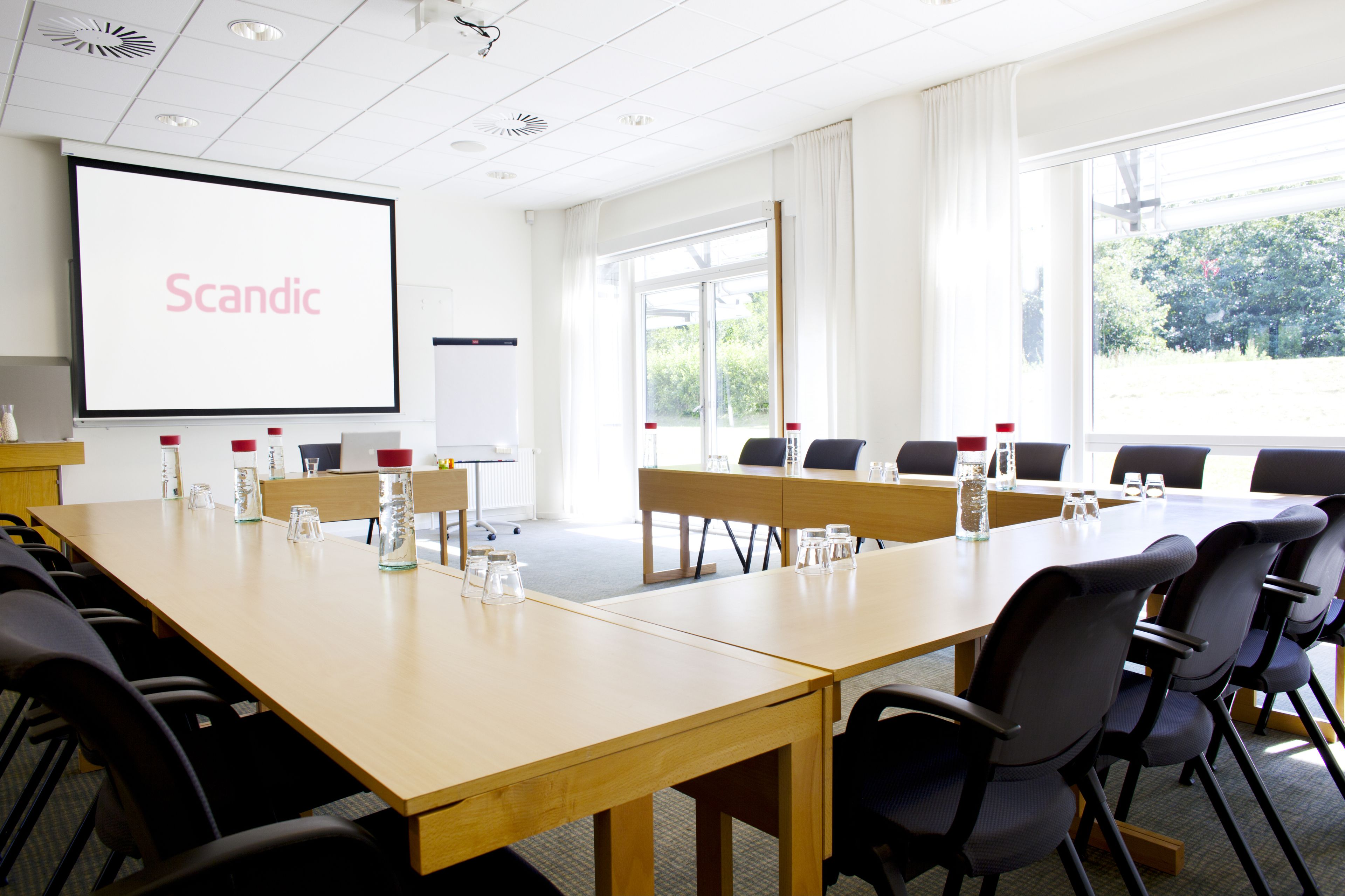 A conference room with a large table, chairs, a projector screen displaying the word "Scandic," and a view of greenery outside.