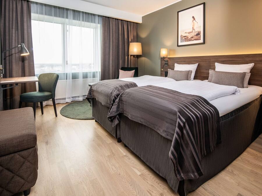 A hotel room with a large bed, a desk, a chair, and a view of the city through the window.