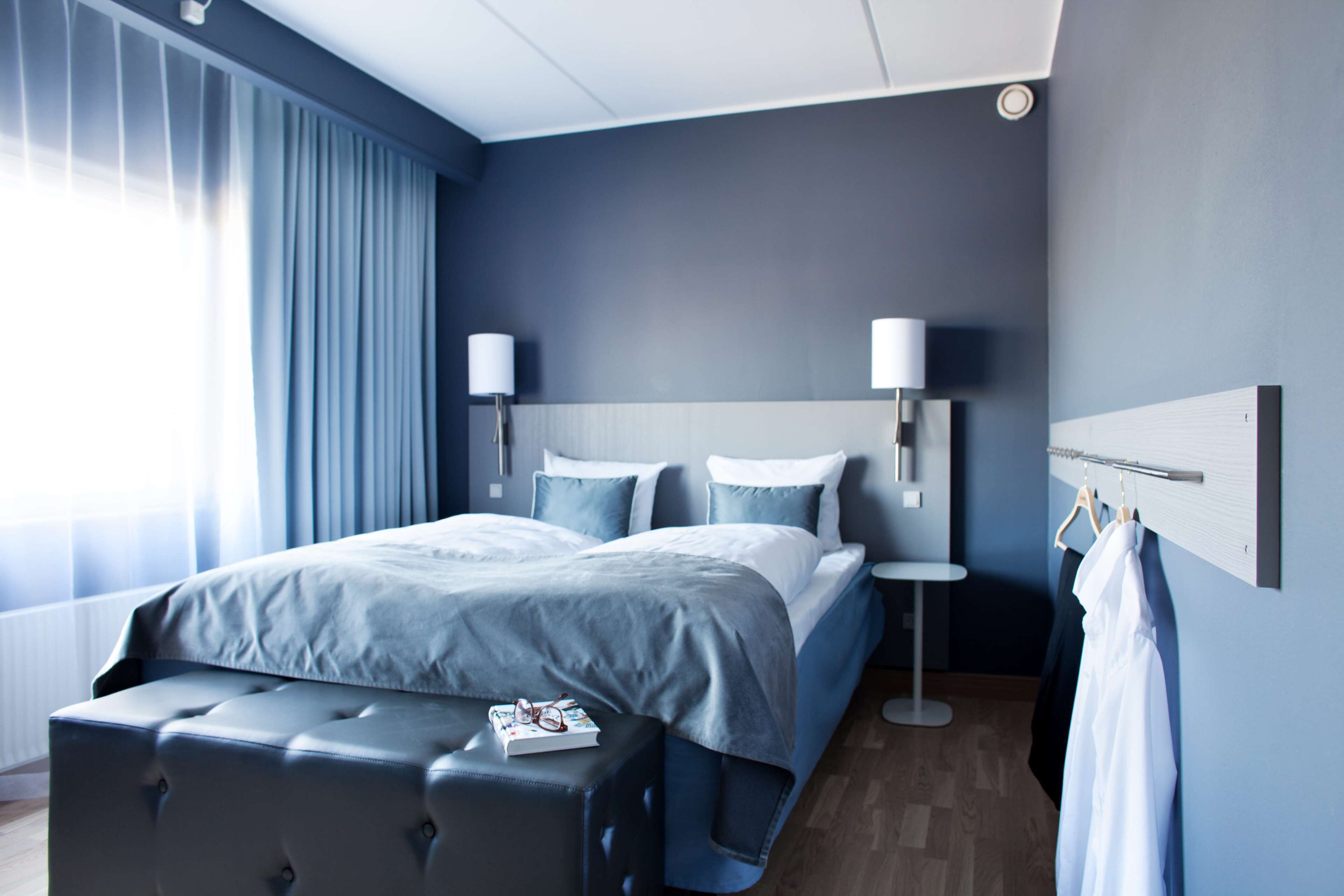 A modern hotel room with a large bed, bedside lamps, a small table, and a hanging coat rack.