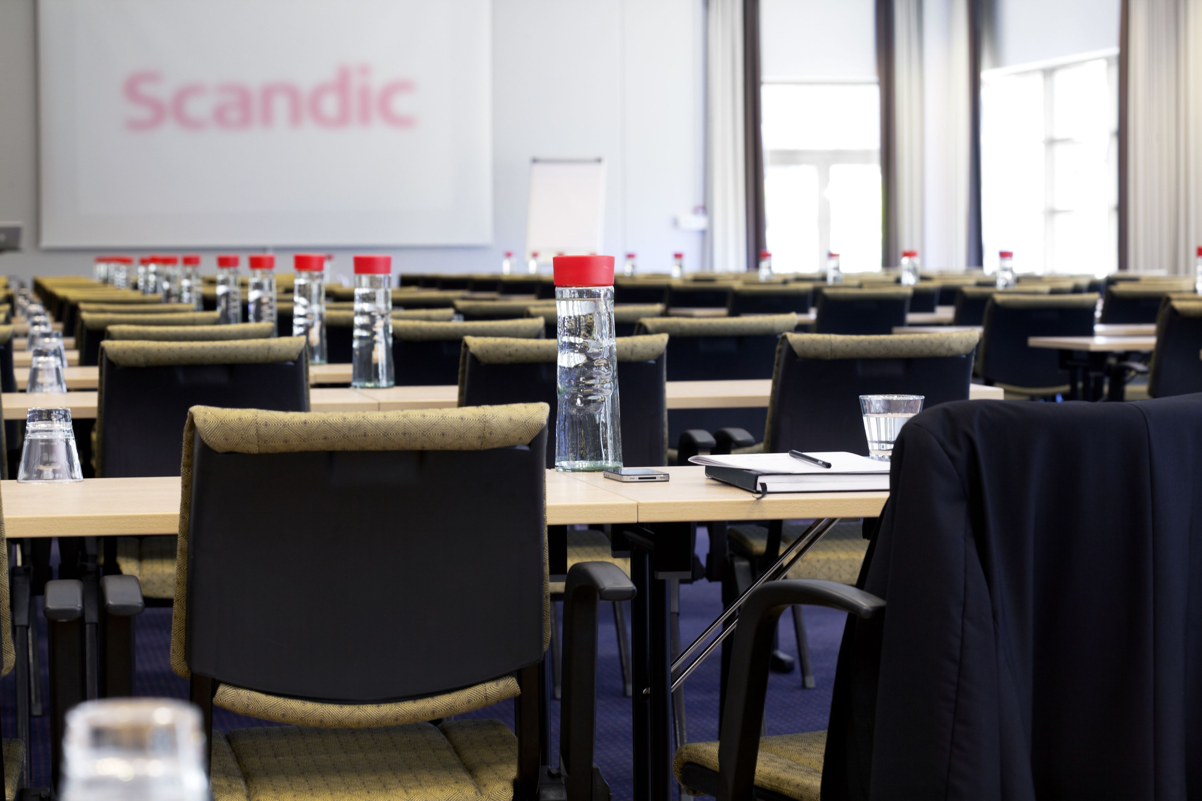 A conference room with rows of tables, chairs, water bottles, and a projector screen displaying the word "Scandic".