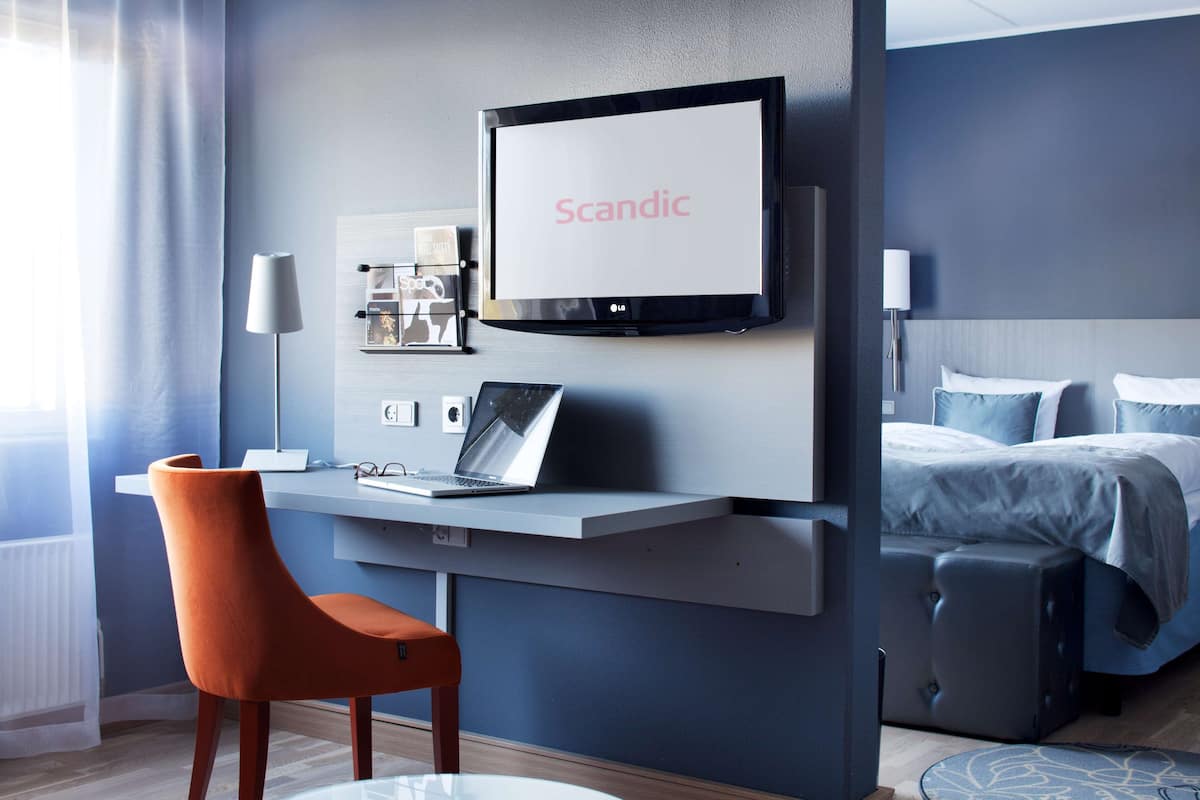 A modern hotel room with a desk, chair, bed, and a TV mounted on the wall displaying "Scandic".