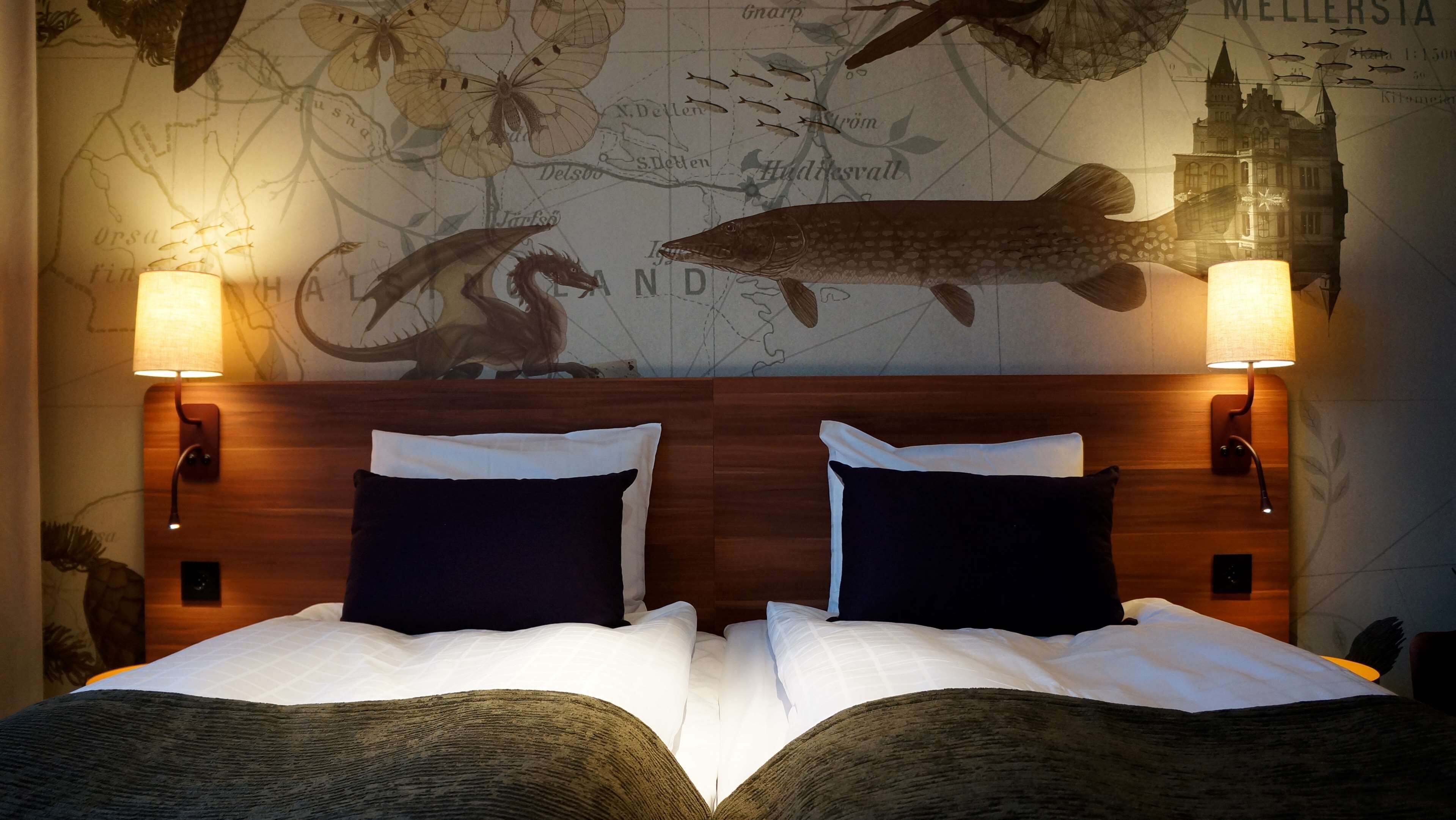 A hotel room with a large map on the wall, two bedside lamps, and a bed with white linens and dark pillows.