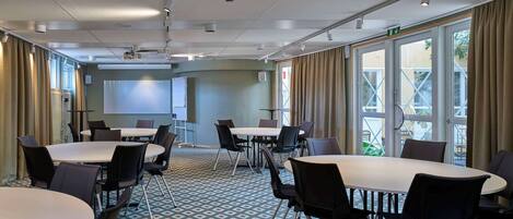 A conference room with tables and chairs, a projector screen, and a large window with curtains.